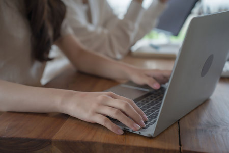 Closeup of businesswomen typing data on keyboard laptop while checking finance graph in document to discussion about investment of new project and enjoying with friendship conversation in workspace.の写真素材