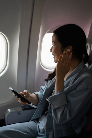 Asian traveler women wear wireless headphones to listening music and typing to chat message on smartphone while sitting near windows during flight business travel in airplane with vacation lifestyle.の写真素材