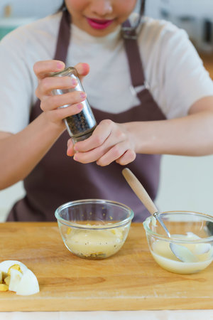 Asian women adding mayonnaise and pepper in chopped boiled eggs to mixing dressing of salad while...
