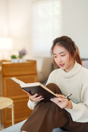 Young asian women in sweater reading the book to learning about knowledge and education while sitting on couch to relaxation and doing activity for spending time with slow life lifestyle at home.の写真素材