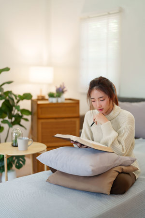 Young asian women in sweater sitting on comfortable couch and reading a book for thinking and learning education while relaxation and doing activity to spending time with slow life lifestyle at home.の写真素材