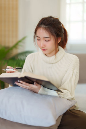 Young asian women in sweater sitting on comfortable couch and reading a book for thinking and learning education while relaxation and doing activity to spending time with slow life lifestyle at home.の写真素材