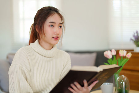 Young asian women in sweater reading a book to thinking and learning about literature story while sitting on couch to relaxation and doing activity to spending time with slow life lifestyle at home.の写真素材