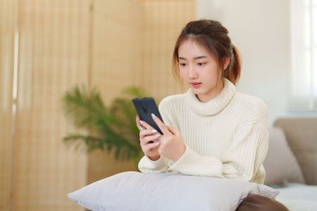 Young asian women in sweater enjoying with surfing entertainment on smartphone while sitting on comfortable couch to relaxation and doing activity for spending time with slow life lifestyle at home.の写真素材