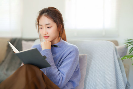 Young asian women in sweater read a book to thoughtful and learning about knowledge education while sitting on sofa to relaxation and doing activity for spending time with slow life lifestyle at home.の写真素材