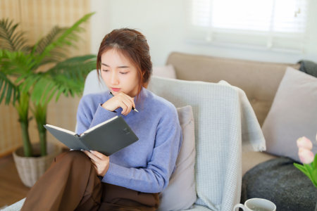 Young asian women in sweater sitting and leaning on couch to reading book and learning about knowledge education while relaxation and doing activity for spending time with slow life lifestyle at home.の写真素材