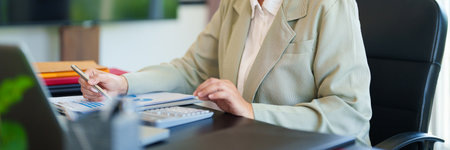 A professional woman in a light suit sits at a desk, focused on her work with documents and a laptop in front of her. The bright office settingの写真素材