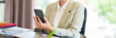 A smiling businesswoman in a light suit is seated at her desk, holding her phone in a bright office...