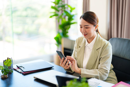 A smiling businesswoman in a light suit is seated at her desk, holding her phone in a bright office setting, looking engaged and connected.の写真素材