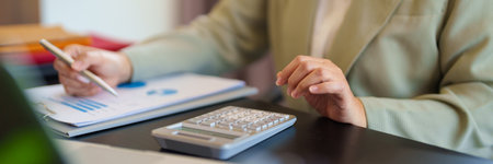 A professional woman in a light suit sits at a desk, focused on her work with documents and a laptop in front of her. The bright office settingの写真素材