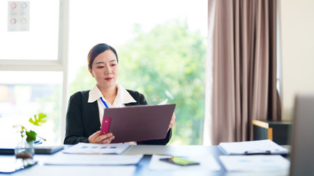 A businesswoman in a black blazer attentively reviews business documents in her office. She sits at her desk in a modern workspace, focused on reading the papers.の写真素材