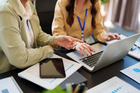 Two businesswomen in formal attire collaborate on a laptop in a bright office. They are both wearing blue lanyards and discussing ideas in a modern workspaceの写真素材