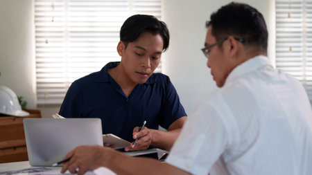 Engineers discussing construction plans at the desk with blueprints and calculators, focusing on project strategies and material estimates.の写真素材