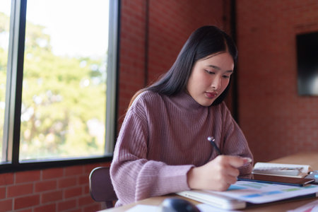A young female designer in a casual sweater is working on design sketches and notes in a modern workspace with a brick wall background. She is focused and engaged in her creative process.の写真素材