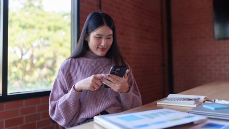 A young female designer in a cozy sweater is using her smartphone while working in a modern studio with natural lighting and a stylish brick wall background.の写真素材