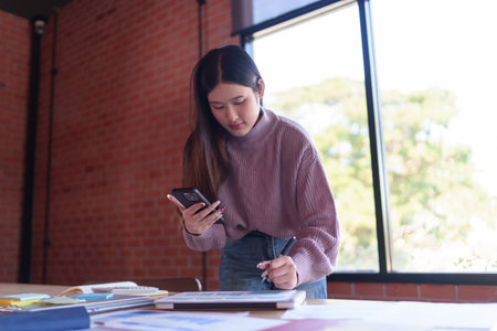 A female designer is reviewing design plans while using her smartphone, standing at a large wooden table filled with creative materials in a modern, well-lit studio.の写真素材