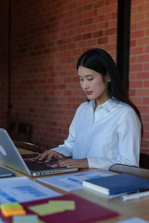 A young businesswoman in a white blouse is working on her laptop at a modern office with an...