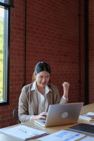 A cheerful businesswoman in a beige blazer raises her fist in excitement while working on her laptop, expressing happiness and success in the workplace.の写真素材