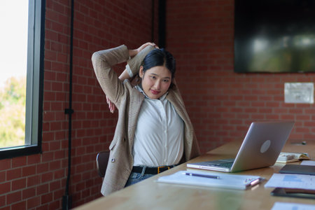A businesswoman in a beige blazer stretches her back while sitting at a wooden desk, feeling muscle fatigue after long hours of work in a modern office with large windows.の写真素材
