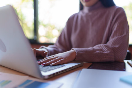 A young female designer in a cozy sweater working on her laptop in a modern office with a brick wall...