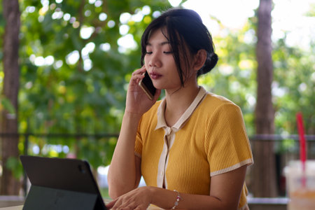 Happy Asian student talking on the phone while working on a tablet in a study areaの写真素材