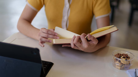Close-up of a woman's hands writing and reading a book on a study desk with a laptop and drink