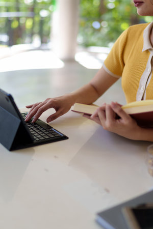 Close-up of a woman's hands writing and reading a book on a study desk with a laptop and drinkの写真素材