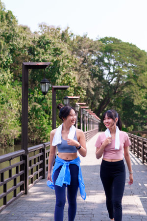 Two young Asian women walking and chatting on a wooden bridge after exercising together in a scenic...