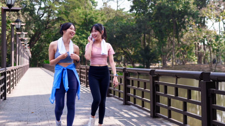 Two young Asian women walking and chatting on a wooden bridge after exercising together in a scenic...