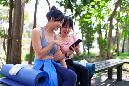 Two young Asian women sitting on a park bench, looking at a smartphone and resting after an outdoor workoutの写真素材