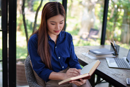 Businesswoman taking notes in a notebook at a natural-themed workspace with laptop, surrounded by...