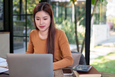 Businesswoman taking notes while working on a laptop in a natural-themed office, showing concentration and efficiency.の写真素材