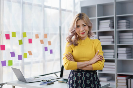 Confident young businesswoman standing with arms crossed in modern office space with sticky notes and laptopの写真素材