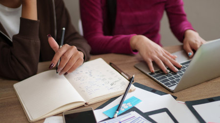 Two adult Asian women UI designers are collaborating on a project, using a notebook, laptop, and wireframes at a desk in a modern office workspace, design concept ideaの写真素材