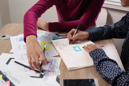 Two adult Asian women are collaborating on a User Interface design, sketching mobile app layouts with pens and paper on a wooden desk. Ideal for UI/UX design content.の写真素材