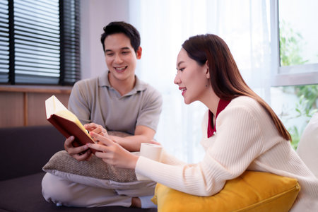 An Asian adult couple is relaxing at home reading a book together. The man and woman are sitting on a sofa with pillows, smiling and sharing story. The woman is holding a cup.の写真素材