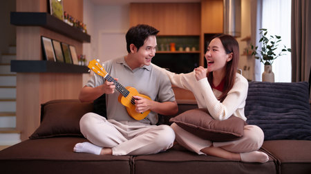 An adult Asian couple is happily spending time together at home. The man plays the ukulele while the woman listens, both smiling and sharing a joyful moment on the sofaの写真素材