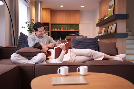 A young Asian couple relaxes at home. The woman is lying on the sofa reading a book while the man is showing her something on his smartphone. Two cups of coffee are placed on table.の写真素材