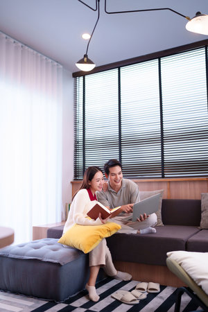 Young Asian couple enjoying leisure time together at home. An adult man is using a laptop while a young woman is reading a book. They are smiling and relaxing indoors promoting happy relationship lifestyle.の写真素材