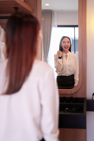 An adult Asian woman smiles at her reflection in a mirror checking her outfit and smartwatch Getting ready for a new day at work or meeting preparation routine.の写真素材