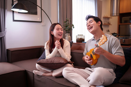 An adult Asian couple is happily enjoying ukulele music in their cozy living room. They are bonding and having a fun leisure time together. Their beautiful relationship is evident in their smilesの写真素材