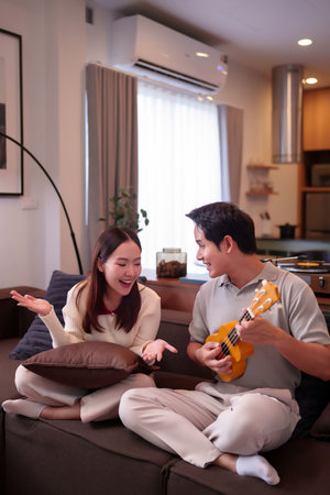 An Asian adult couple enjoys playing ukulele on sofa. Man is playing instrument while woman is laughing. Lovely time at home with musical instrument ukulele.の写真素材