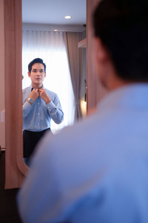 Asian adult man adjusting his bow tie in the mirror getting ready for a special event preparing for a celebration wedding concept perfecting his look wearing formal dress codeの写真素材