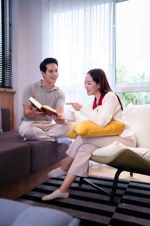 Asian couple enjoying reading book together at home. Asian woman relaxing on chair and adult man reading book smiling happy. They are spend time together cozy living roomの写真素材