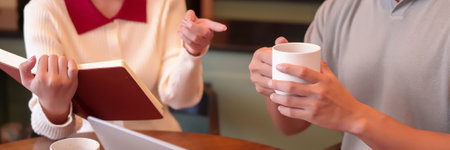Asian adult woman showing notebook to a man holding a cup in a cafe. Business discussion,...