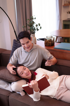 An Asian adult couple is relaxing on a brown sofa in their living room. The woman is reading a book while lying down, with her head on a pillow. The man is sitting up, holding a smartphone and smiling. They appear happy and content enjoying a relaxing moment at home.の写真素材