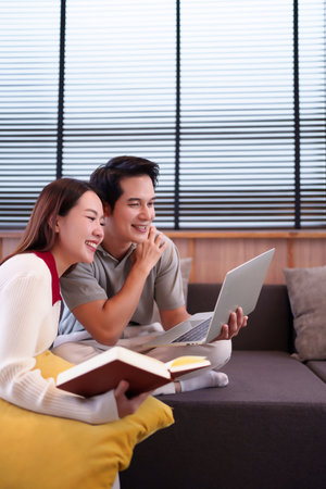 A happy Asian couple, an adult man and woman, are smiling and relaxing together at home on the sofa. The man holds a laptop while the woman reads a book, enjoying their leisure time.の写真素材