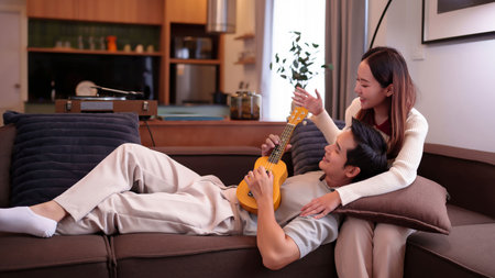 Asian young couple resting on the sofa at home. The man is playing ukulele while the woman is enjoying the music. Sweet moment, romantic atmosphere, happy together at home.の写真素材