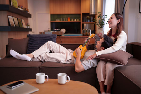 Capture a heartwarming moment of an Asian adult couple enjoying leisure time at home. He plays ukulele, while she laughs relaxing on the sofa creating cherished memories together.の写真素材