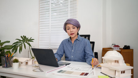 An Asian women architect plans and designs with a laptop on a desk. The architect is working on an architecture project, creating building and landscape blueprints with precision and skill in officeの写真素材